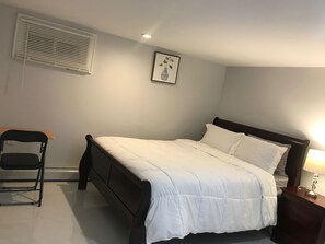 A bedroom with a bed, a chair, a bedside table with a lamp, a wall-mounted picture, and a window with blinds. - We have beautiful, serene environment for your comfort. (Queens)