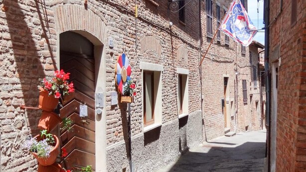 Une rue étroite bordée d’immeubles en briques, de pots en terre cuite ornés de fleurs et d’un drapeau.