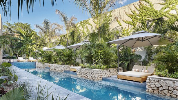 Un área junto a la piscina con sillones y sombrillas, rodeada de abundante vegetación y muros de piedra.