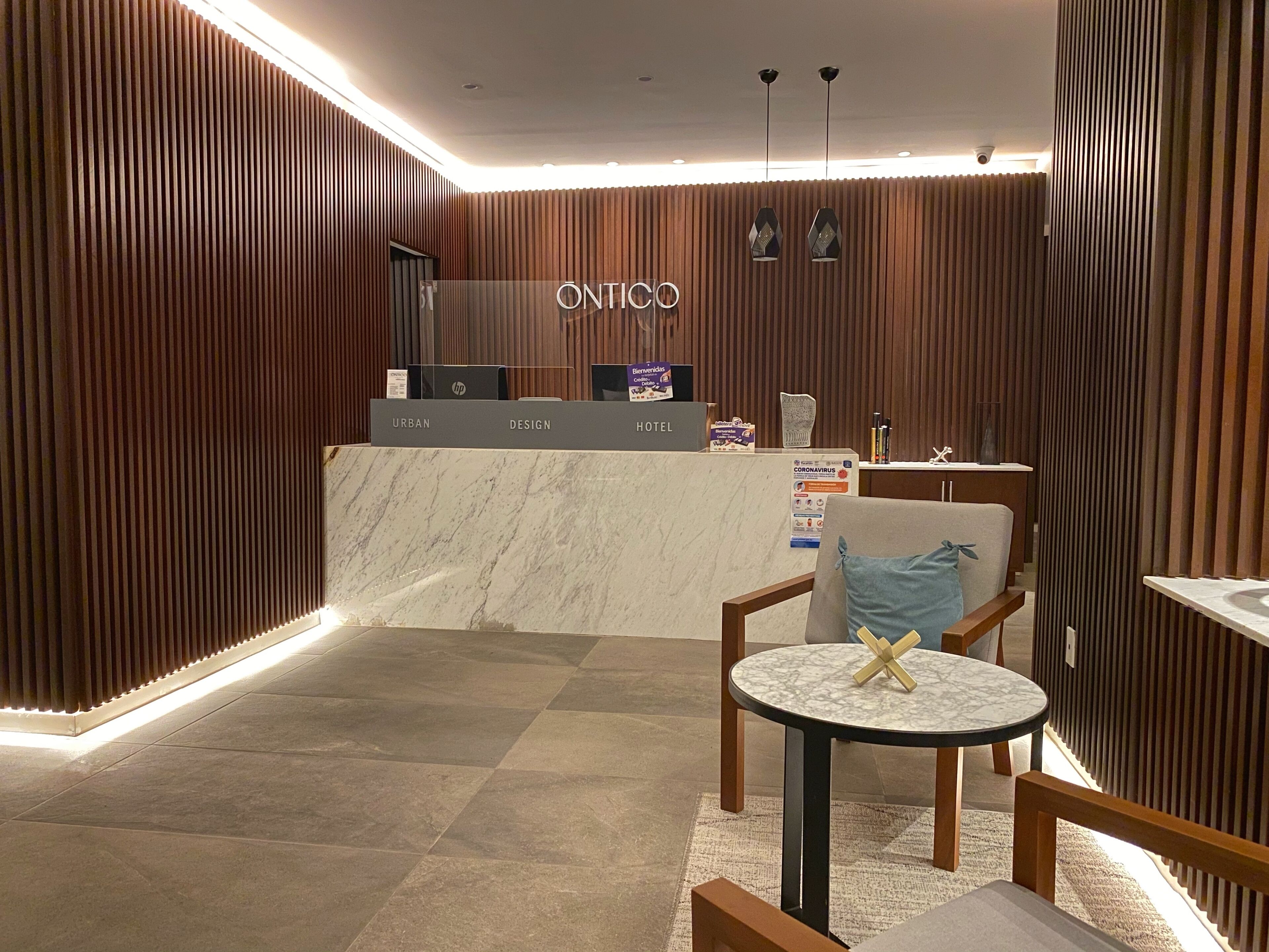 A hotel reception area with a wooden panel wall, a reception desk labeled "ONTICO," and a small round table with a chair.