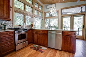 Une cuisine dotée de meubles en bois, d’une cuisinière en acier inoxydable, d’un lave-vaisselle et de grandes fenêtres offrant une vue sur les arbres.