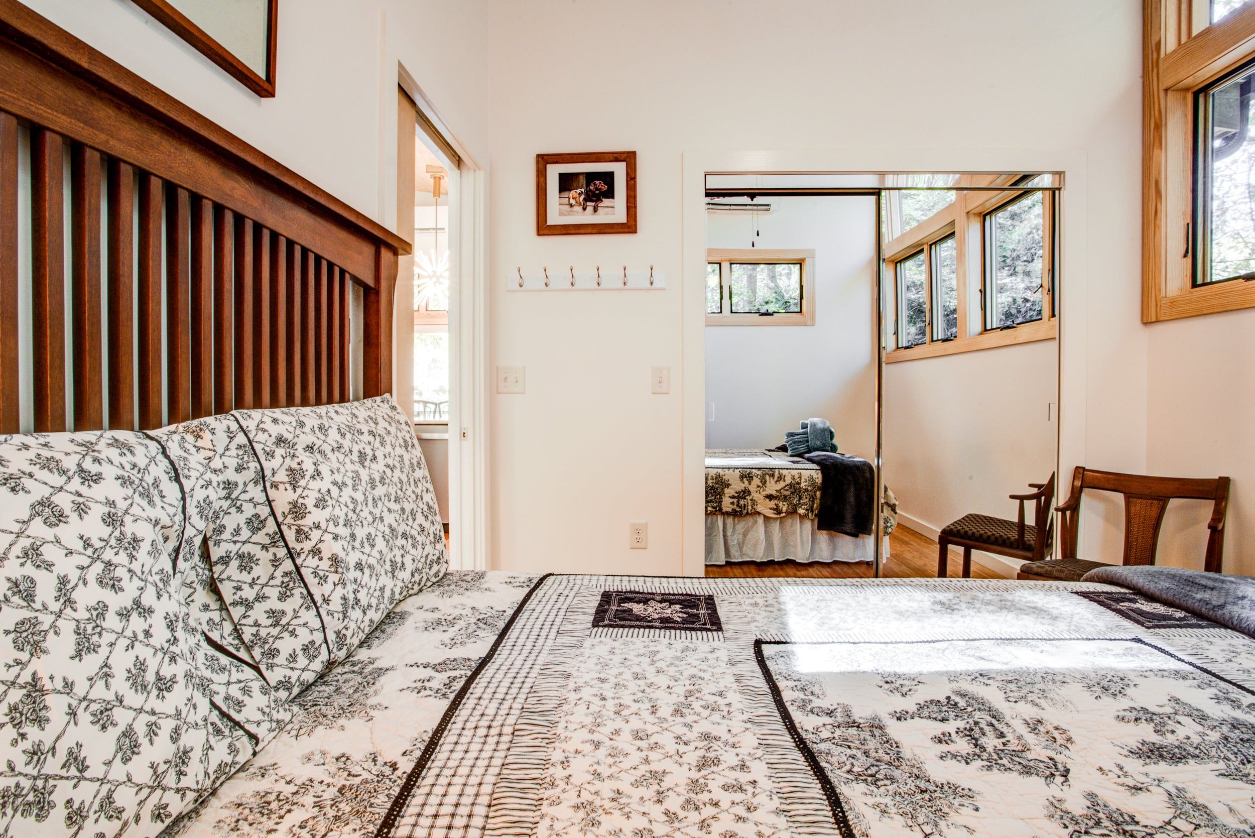 Un dormitorio con cama, cabecera de madera, mesita de noche, una silla, ventana y puerta que da a otra habitación.