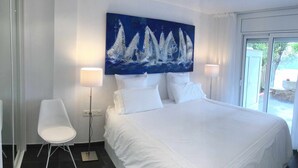 Hotellihuone, jossa on suuri sänky, valkoiset vuodevaatteet, seinällä purjeveneaiheinen taulu, kaksi yöpöytälamppua ja näkymä lasiovesta.