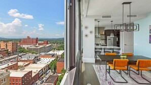 A modern apartment with a large balcony, a dining area with orange chairs, and a city view. - Endless Sunset Retreat | Arras Vacation Rentals (Asheville)