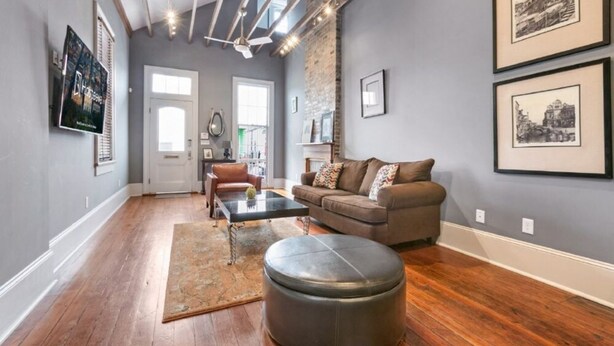 Un salon doté d’un parquet, d’un canapé en cuir marron, d’une table basse et d’un pouf noir. Les murs sont ornés de tableaux encadrés et une porte donne accès à une autre pièce.
