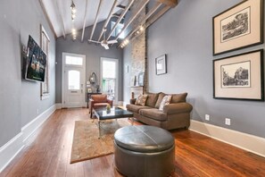 Un salon doté d’un parquet, d’un canapé en cuir marron, d’une table basse et d’un pouf noir. Les murs sont ornés de tableaux encadrés et une porte donne accès à une autre pièce.