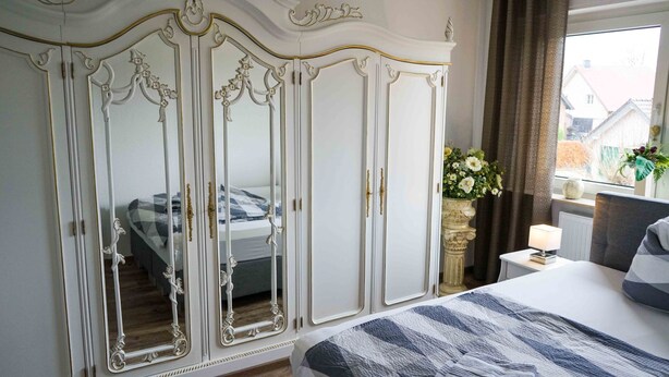 A bedroom with a large white wardrobe, a bed with a striped blanket, a bedside table with a lamp, and a window with curtains. - Ferienwohnung Anke-Apartement 5b (Heinsberg)