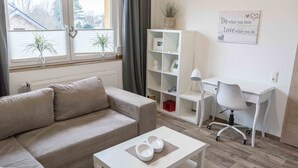 A modern living room with a sofa, a coffee table with pet bowls, a white desk with a chair, and a bookshelf. - Ferienwohnung Anke-Apartement 5d (Heinsberg)