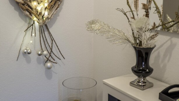 A bathroom with a decorative wall hanging, a vase with dried plants, a glass container with moss, and a mirror on the vanity. - Ferienwohnung Anke-Apartement 5d (Heinsberg)