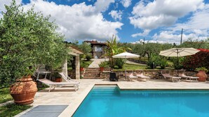 Une piscine entourée de verdure, avec des chaises longues et des parasols, et un chemin de pierre qui mène à un bâtiment.