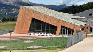 Un edificio moderno con tejado inclinado y amplios ventanales de vidrio, rodeado por una valla metálica y un muro de piedra.