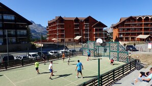 Una cancha de baloncesto al aire libre con jugadores, rodeada de edificios de madera y montañas al fondo.
