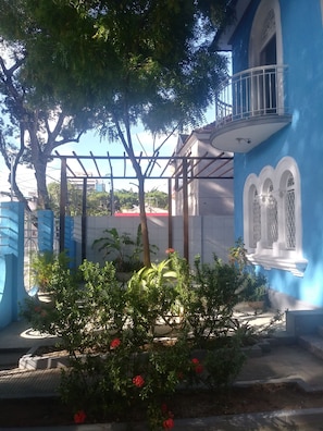 Un bâtiment bleu doté d’un balcon, d’une pergola et d’un jardin agrémenté de fleurs rouges.