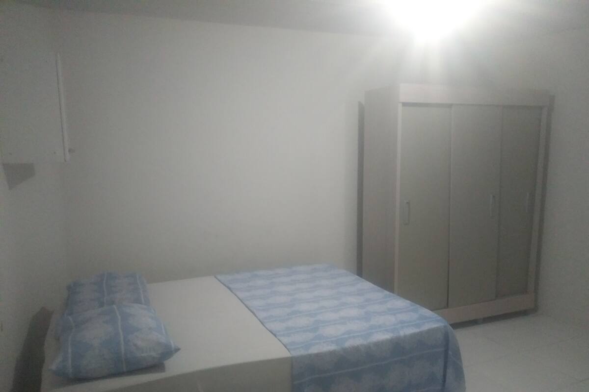 A small, dimly lit room with a single bed, a wardrobe, and a wall-mounted light.