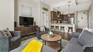 Ein modernes Wohnzimmer mit Flachbildfernseher, grauen Sofas, einem Couchtisch und einer Küchenzeile mit Barhocker.