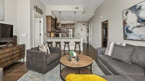 Ein modernes Wohnzimmer mit einer Couch, einem Couchtisch und einem Fernseher. Es gibt eine Küchenzeile mit einem Kühlschrank und Barhocker.