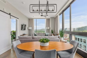 Een moderne woonkamer met een ronde houten tafel, grijze banken en een groot raam met uitzicht op de stad.