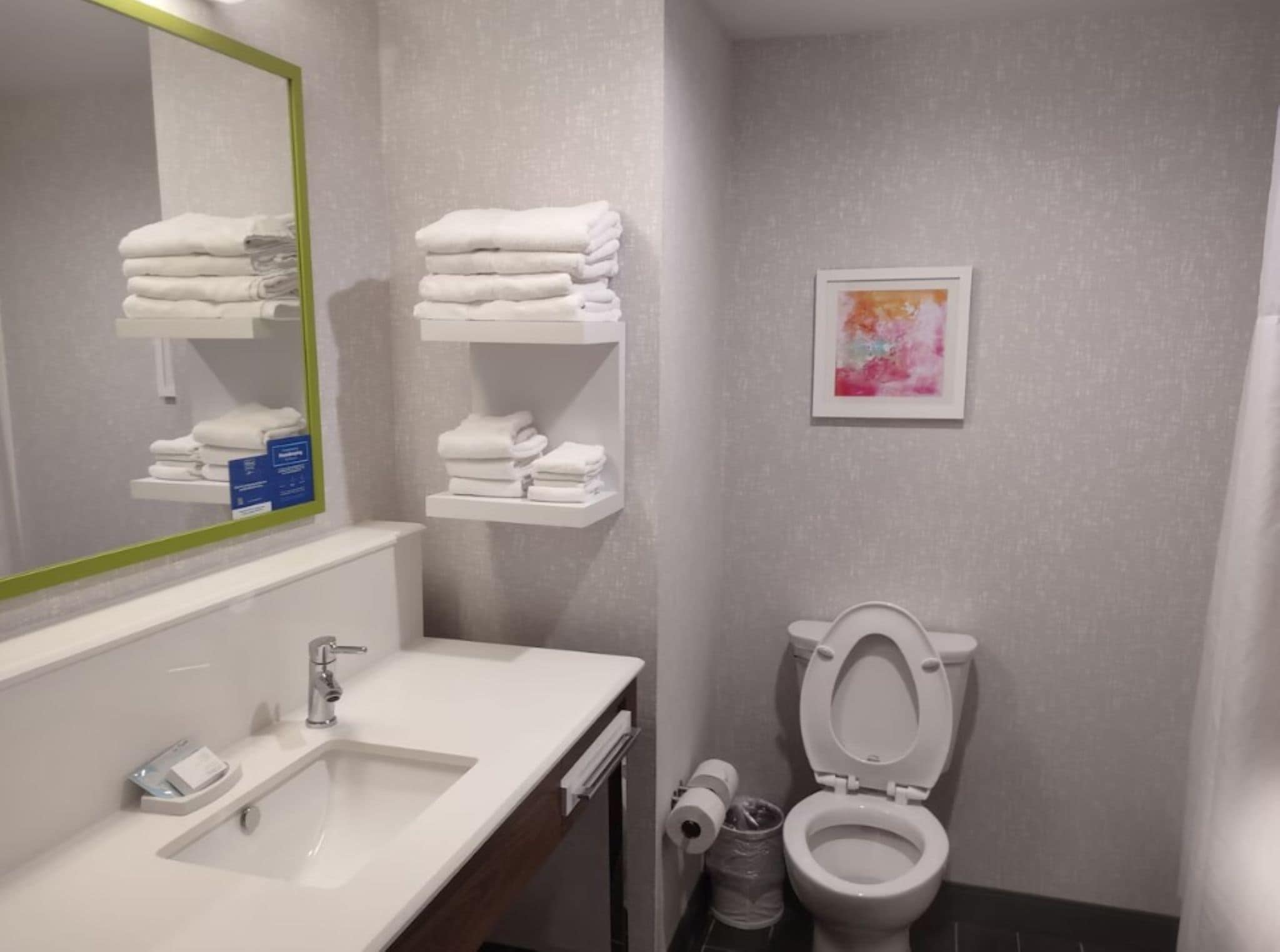 Un baño con lavamanos blanco, espejo con marco verde, inodoro y repisas con toallas dobladas.