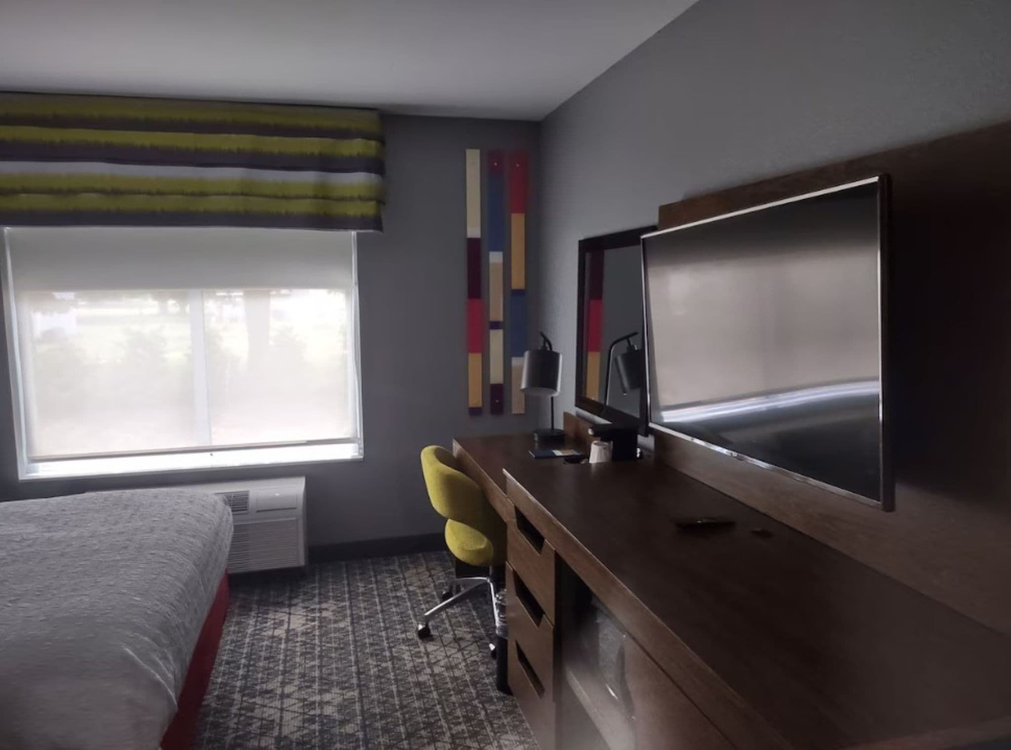 A hotel room with a bed, a desk with a chair, a large TV, and a window with blinds.