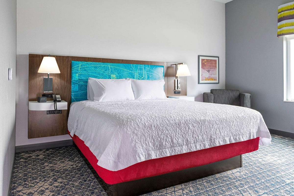 A hotel room with a large bed, two bedside lamps, a headboard with a cityscape design, a red accent on the bed, and a patterned carpet.