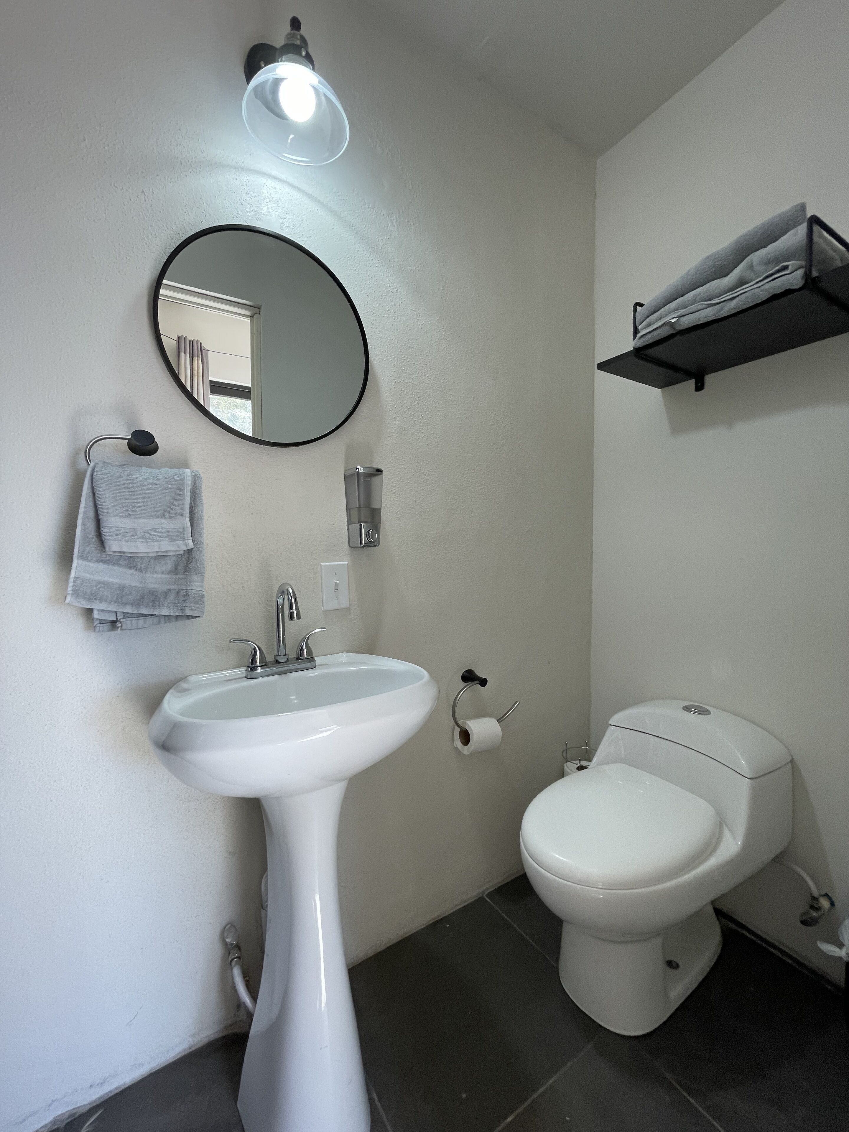 Un baño con lavamanos blanco, un espejo redondo, inodoro y toallero.