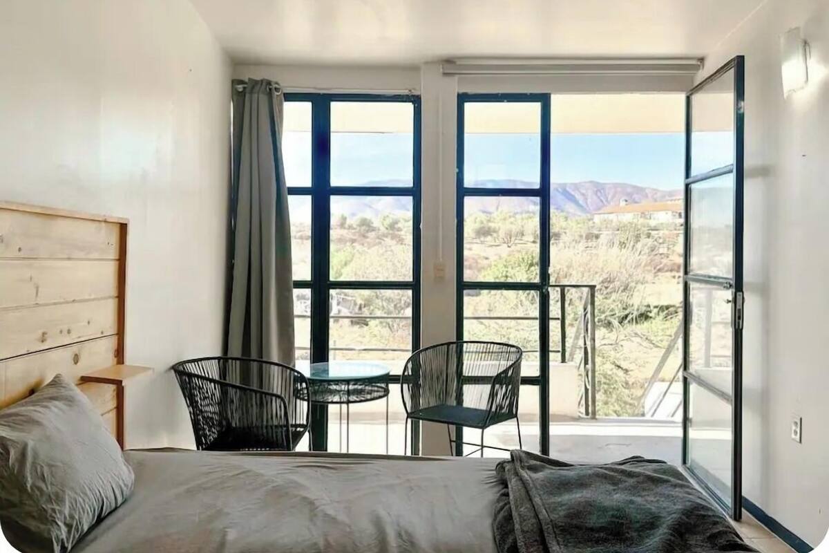 A bedroom with a bed, two chairs, a small table, and large windows offering a view of a desert landscape.