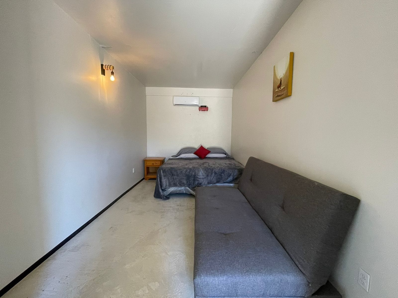 A small, narrow room with a grey sofa, a bed with grey bedding, a small wooden nightstand, and a wall-mounted air conditioner.