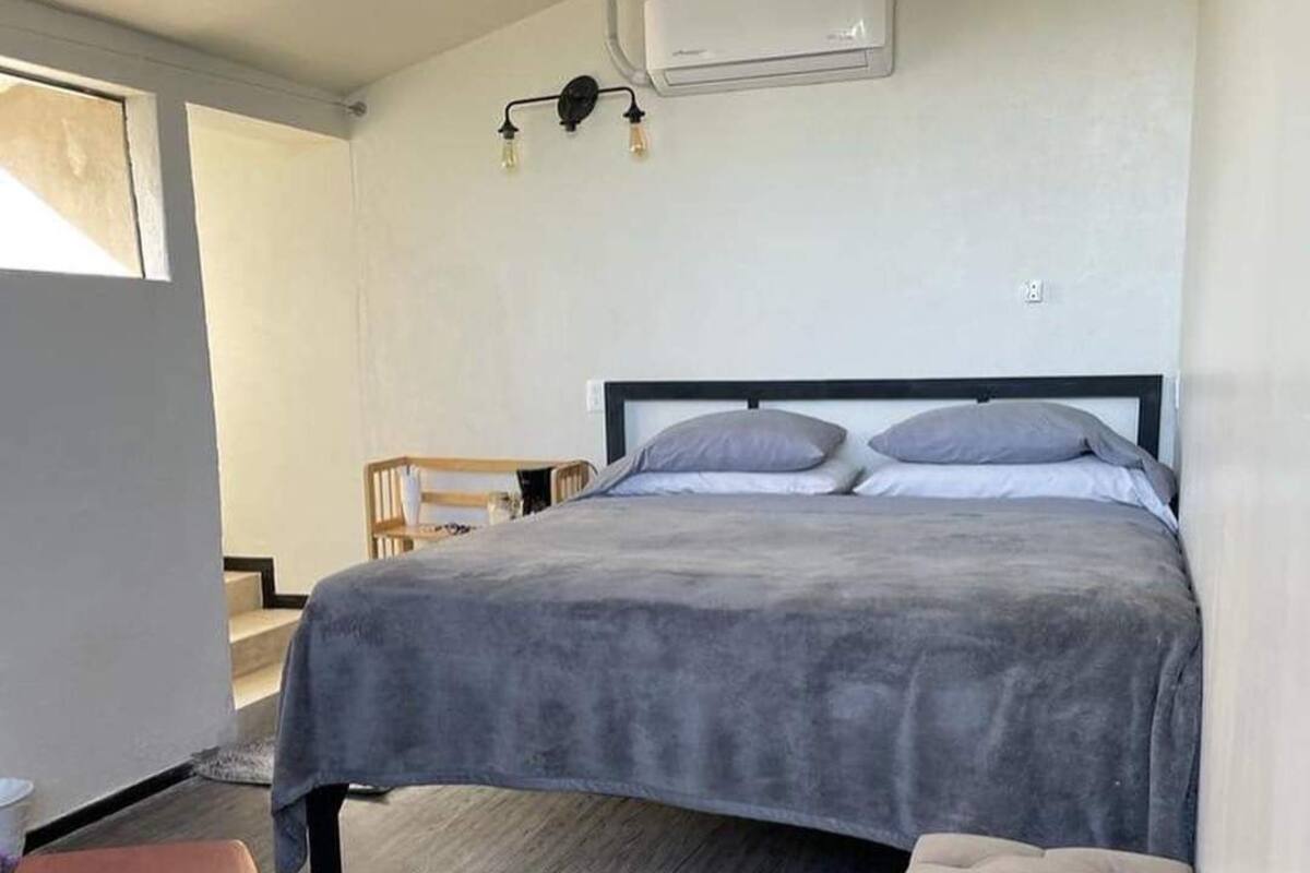 A bedroom with a bed, a bedside table, a chair, a bench, and an air conditioning unit on the wall.