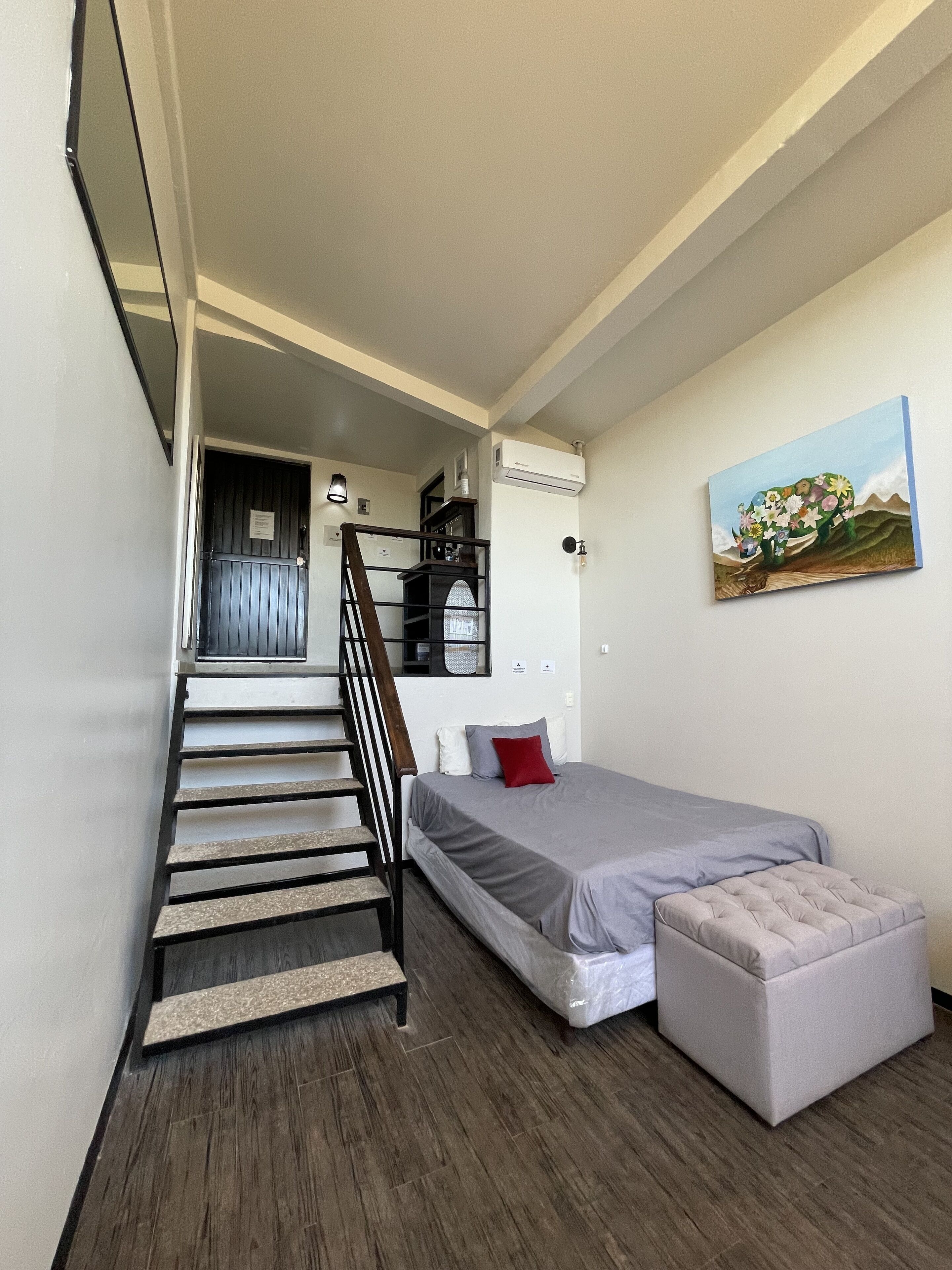 A bedroom with a bed, a small stool, a staircase, and a painting on the wall.
