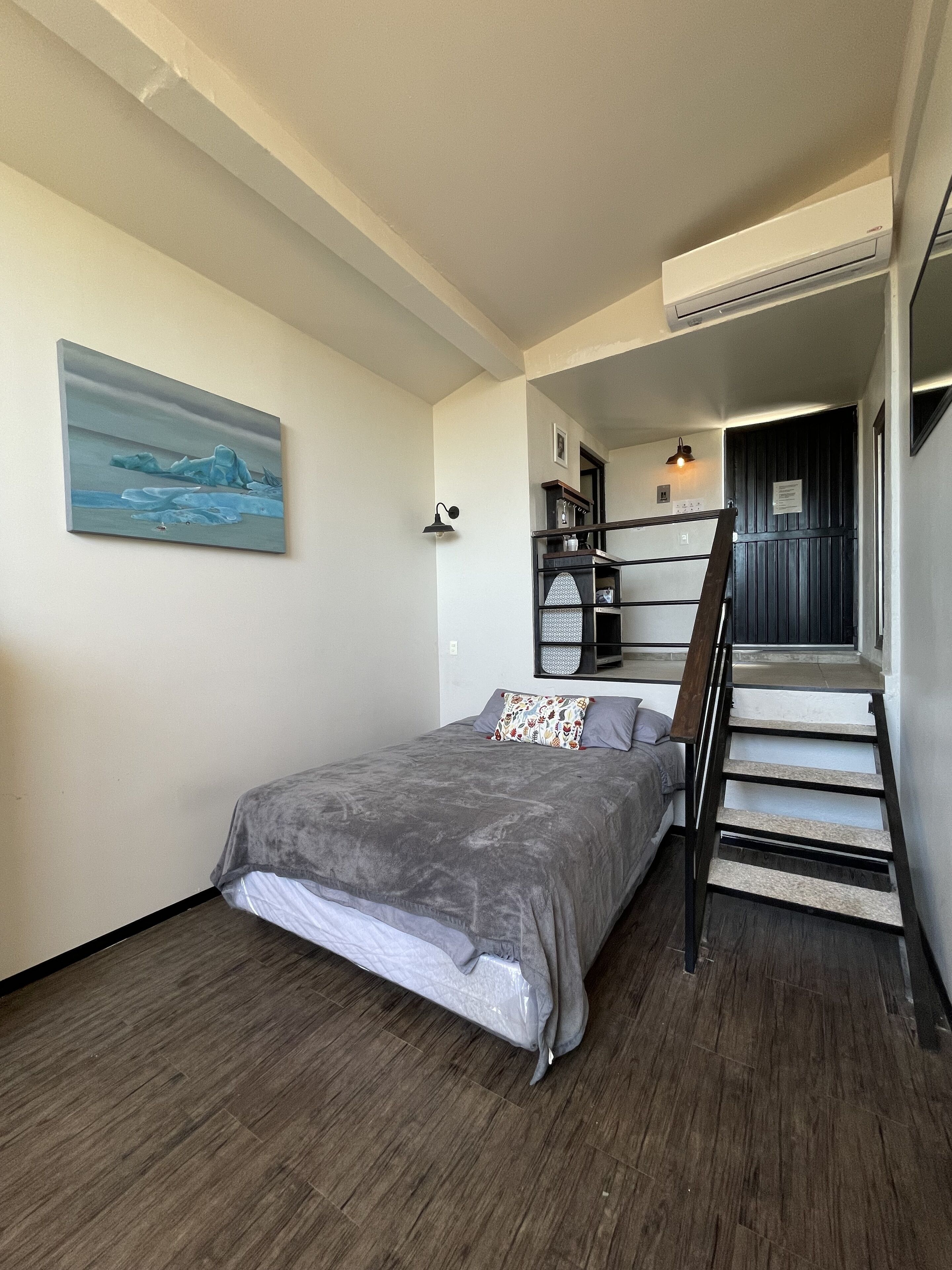 Quarto com cama, escada, quadro na parede e pequena cozinha.