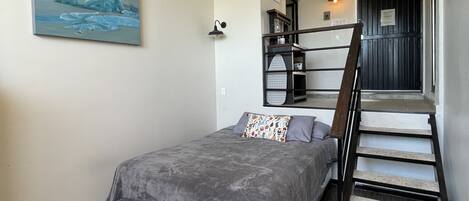 Quarto com cama, escada, quadro na parede e pequena cozinha.