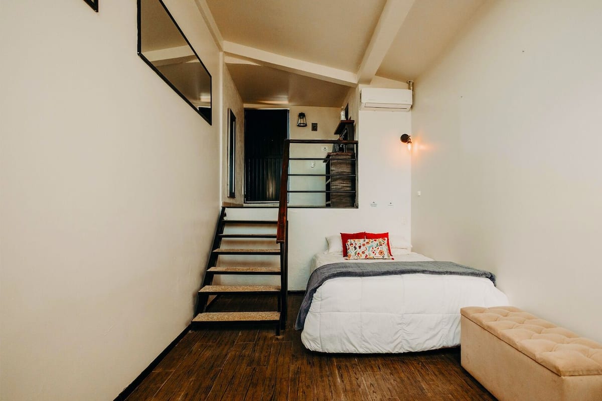 A compact bedroom with a bed, a bench, and a staircase leading to another room.