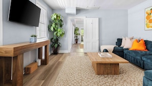 A living room with a wooden coffee table, a blue sofa with orange pillows, a TV mounted on the wall, and a ceiling fan. - Historic Charleston Style House (Charleston)