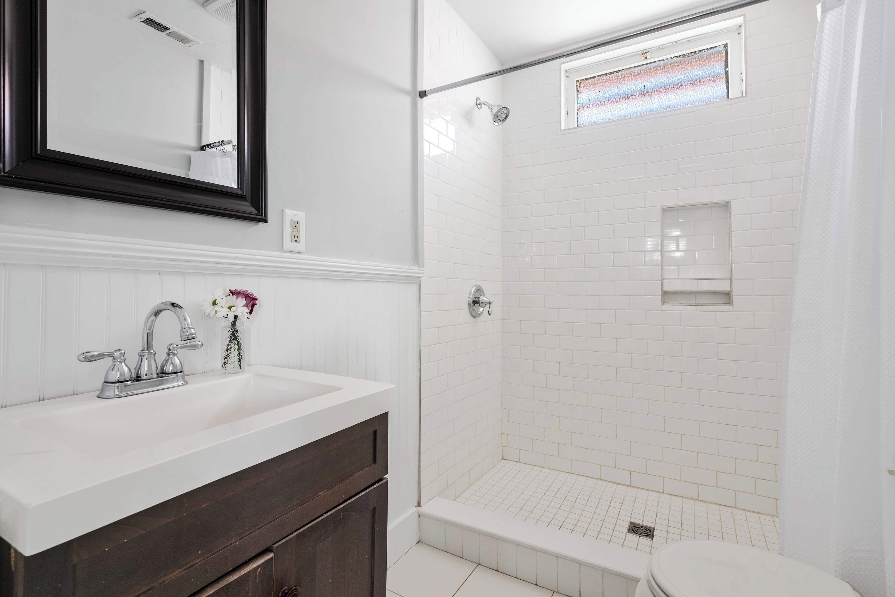 A bathroom with a white sink, dark wooden vanity, a shower with white tiles, and a small window above the shower.