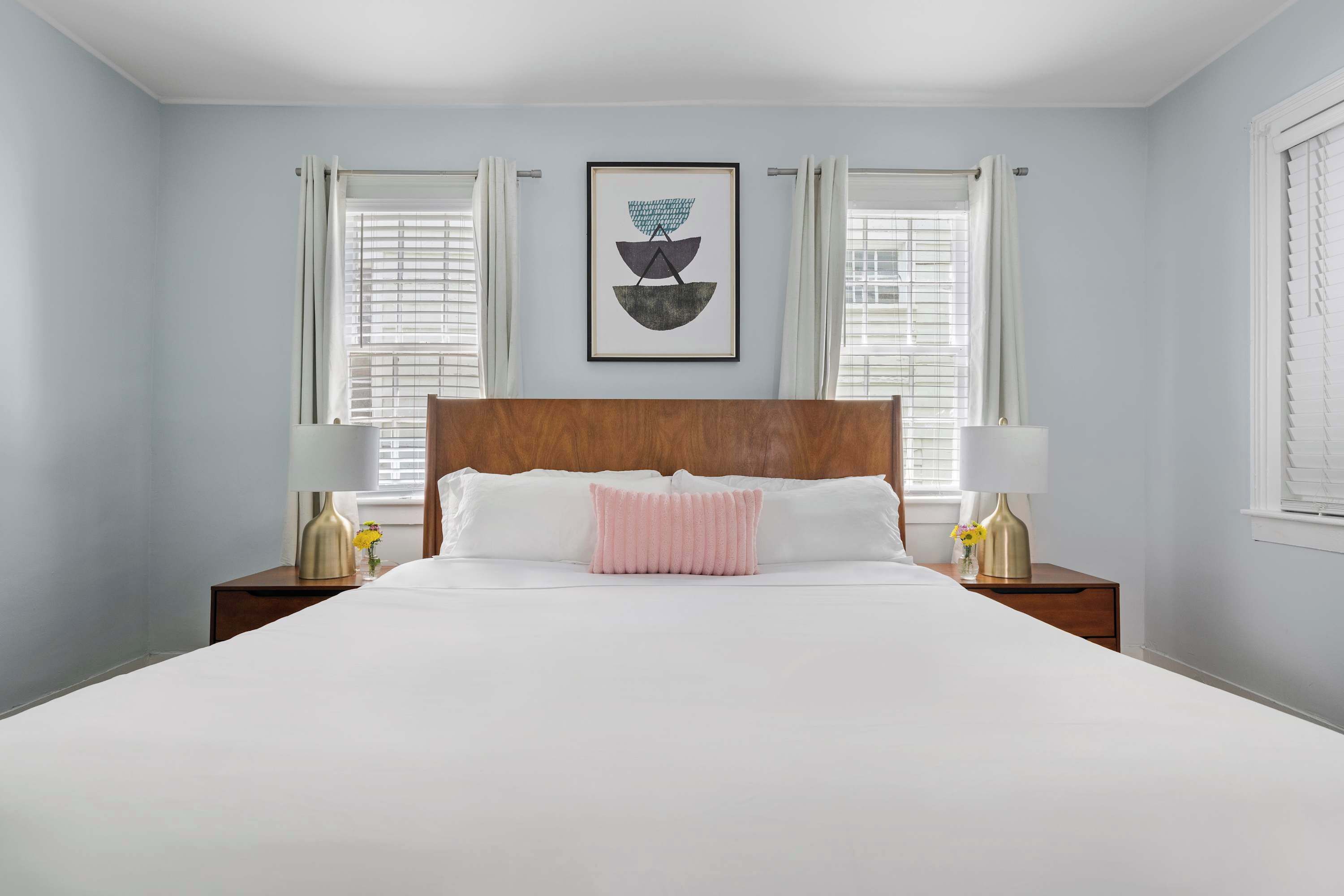 A neatly made bed with white linens and a pink accent pillow, flanked by two bedside tables with lamps and vases.