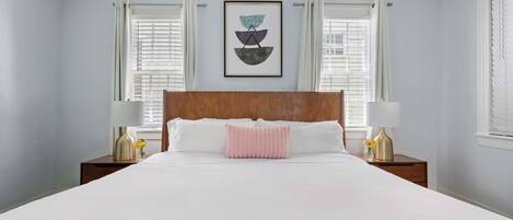 A neatly made bed with white linens and a pink accent pillow, flanked by two bedside tables with lamps and vases.