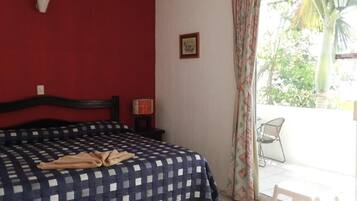A bedroom with a checkered bedspread, a single bed, a nightstand with a lamp, a framed picture on the wall, and a view of a patio with a chair and a palm tree.