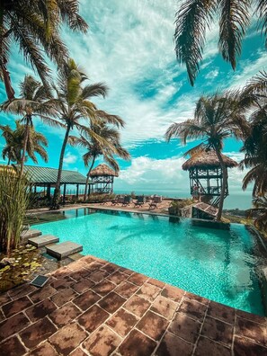 A pool area with clear blue water, palm trees, and thatched-roof structures. - Hacienda Cocuyo (Samaná)
