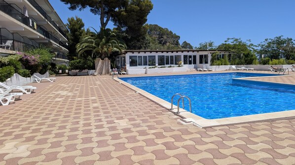 Ein Schwimmbecken mit Fliesen umrandet, Liegestühle und ein Gebäude mit großen Fenstern im Hintergrund.