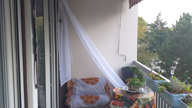 A balcony with a covered seating area, a small table with a colorful tablecloth, and potted plants. - The secret of good taste. (Vaulx-en-Velin)