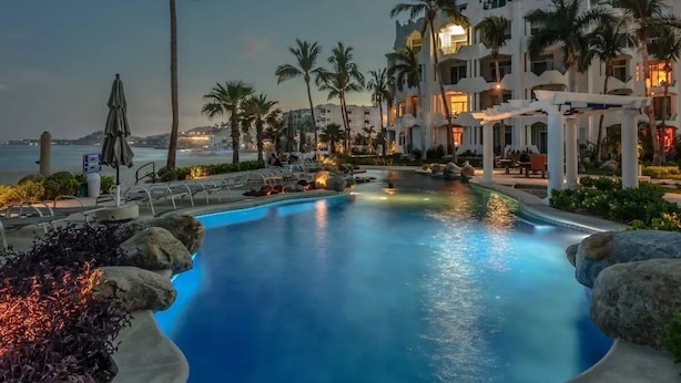 A resort pool with palm trees and a beach view, surrounded by white buildings with balconies.