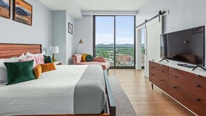 Une chambre moderne avec un grand lit, une télévision, une commode et une porte coulissante donnant sur un balcon avec vue sur la ville.