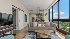 Ein modernes Wohnzimmer mit Sofa, Couchtisch und Fernseher, großen Fenstern mit Blick auf die Stadt und einer Küche im Hintergrund.