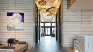 Eine moderne Hotelhalle mit einer langen Rezeption, einer Sitzecke mit einer Couch und Sesseln, einem großen abstrakten Gemälde und einer Decke mit leuchten blattformigen Lampen.