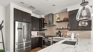 A modern kitchen with dark cabinets, stainless steel appliances, a marble countertop, and a pendant light.