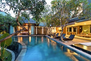 A modern outdoor pool area with lounge chairs, a wooden deck, and a building with large windows. - Paya Paya Villa (Seminyak)