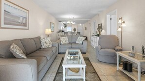 Una sala de estar con sofá, sillón y mesa de centro, un comedor con mesa y sillas, y una cocina al fondo.