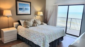 A hotel room with a bed, two bedside lamps, a nightstand, a framed picture on the wall, and a view of the beach through a sliding door.