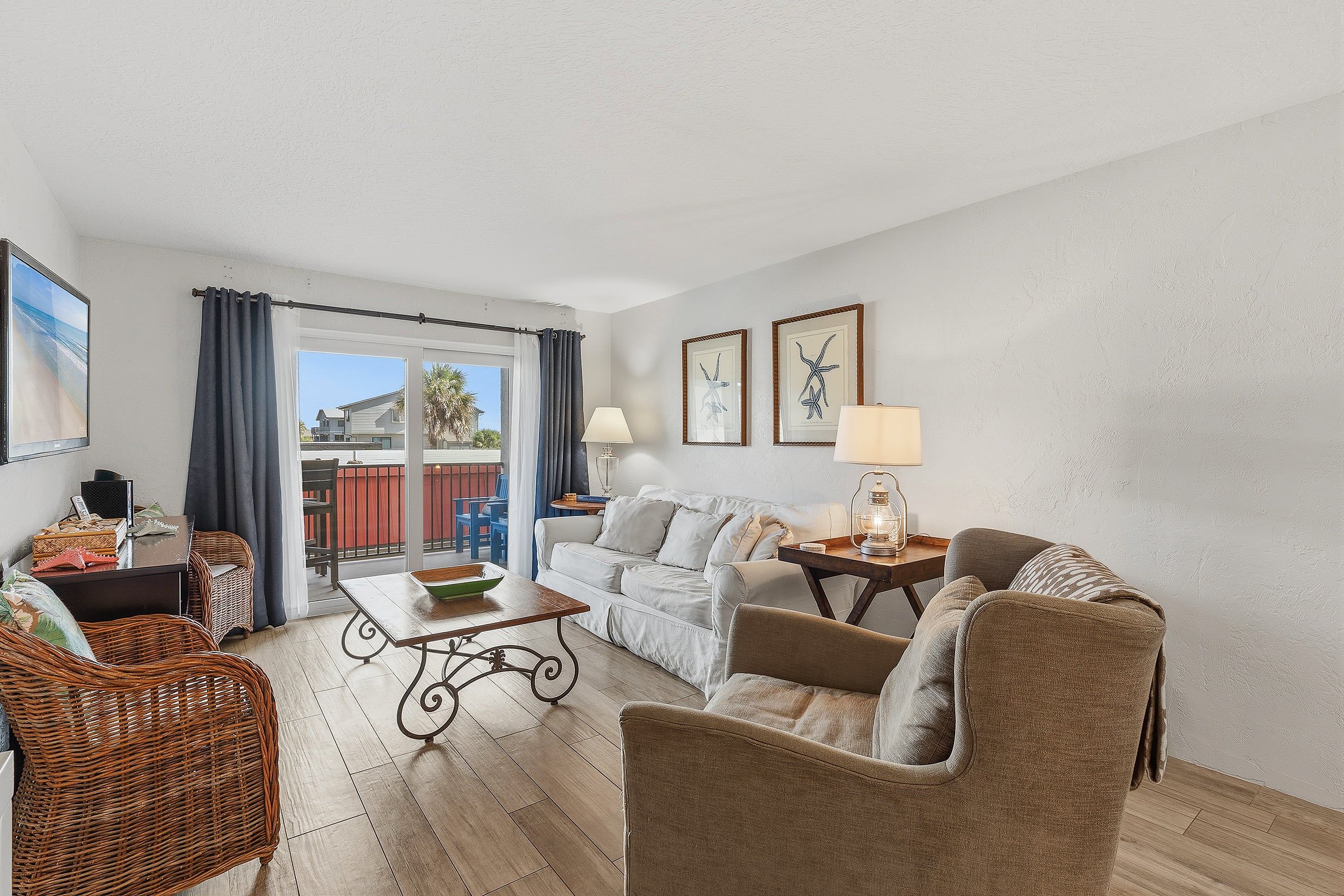 Un salon comprenant un canapé, des fauteuils, une table basse et une table à manger. On y trouve deux lampes, un fauteuil en osier et, à travers une porte vitrée, une vue sur l’extérieur.