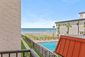 A poolside view with a beach and ocean in the background, a fenced area with lounge chairs, and a building with a flat roof. - 206 - Ocean View 2 bedroom (New Smyrna Beach)